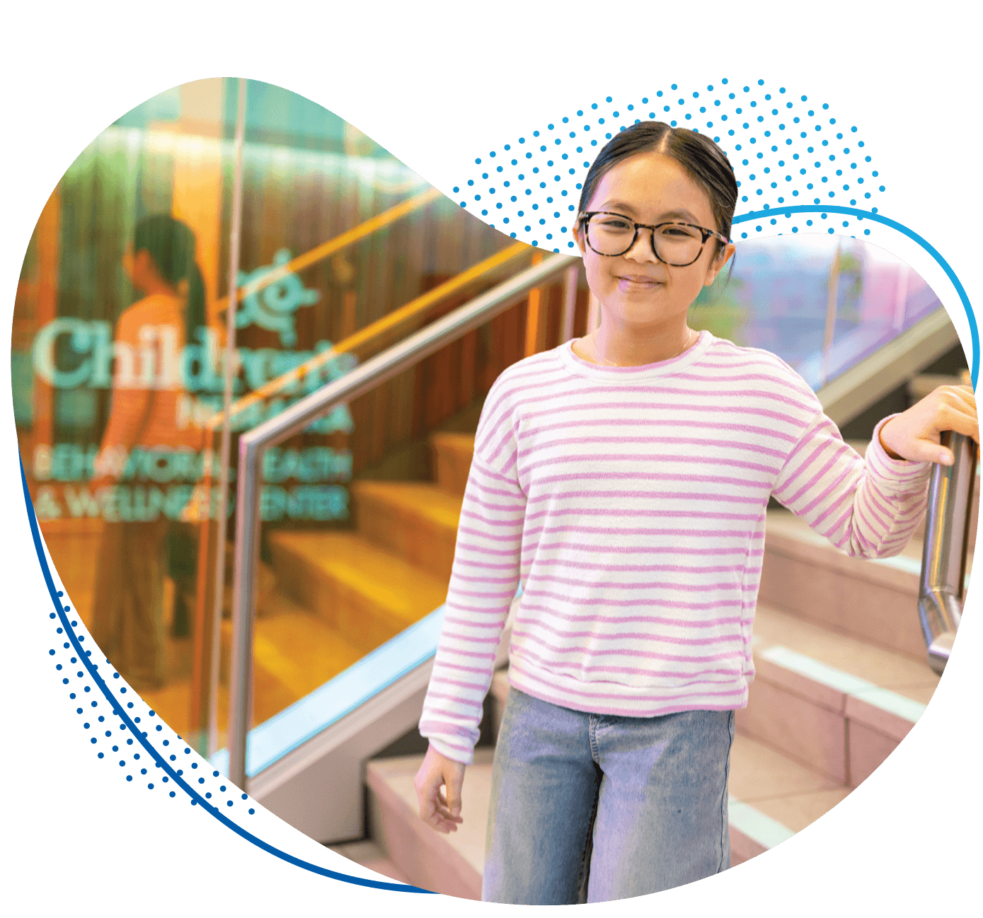 Young girl in pink striped sweater and glasses standing in the entrance of the new Behavioral Health & Wellness Center at Children's Nebraska.
