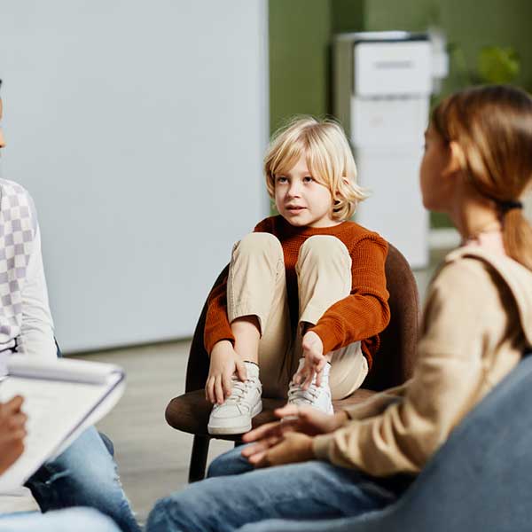 Young child in rust-colored sweater sitting in chair with knees pulled up, surrounded by people in a classroom setting.