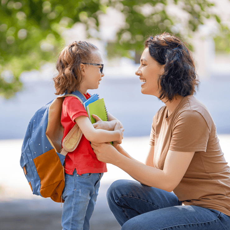 Parent talking to young child about school under a large shade tree, in a bright outdoor park setting.