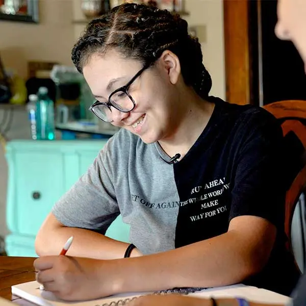 Lilly smiling while writing in a notebook at a table in a home setting.